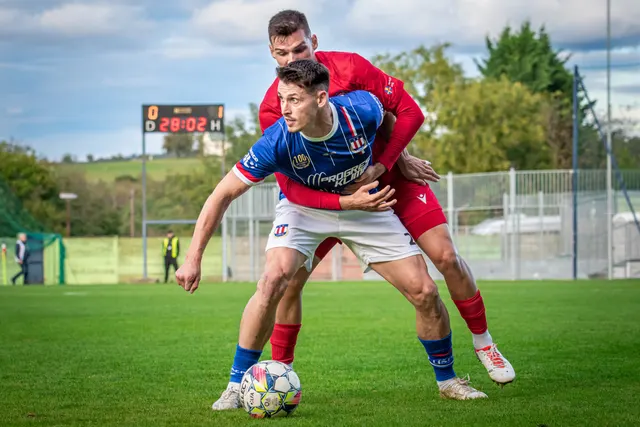 Fotbalista v modrém dresu si kryje míč před soupeřem v červeném